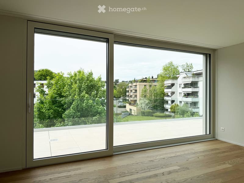 Empty room with wooden flooring, large sliding glass doors leading to a balcony with a view of greenery and buildings.