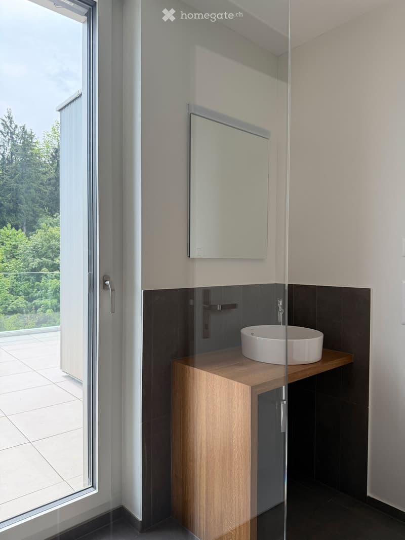 Modern bathroom with a wooden sink cabinet, white sink, and a large mirror. The room features dark tiles and a large window offering a view of greenery outside.