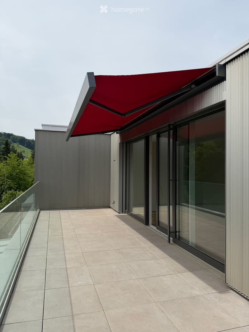 Modern balcony with a red awning, glass railing, and tiled floor, overlooking a green landscape.