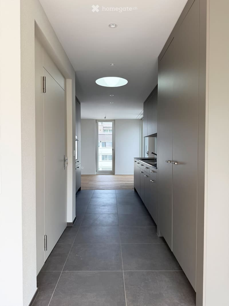 Modern hallway with grey tiled floor, white walls, built-in cabinets, and recessed lighting. A doorway leads to a room with a view of the outside.