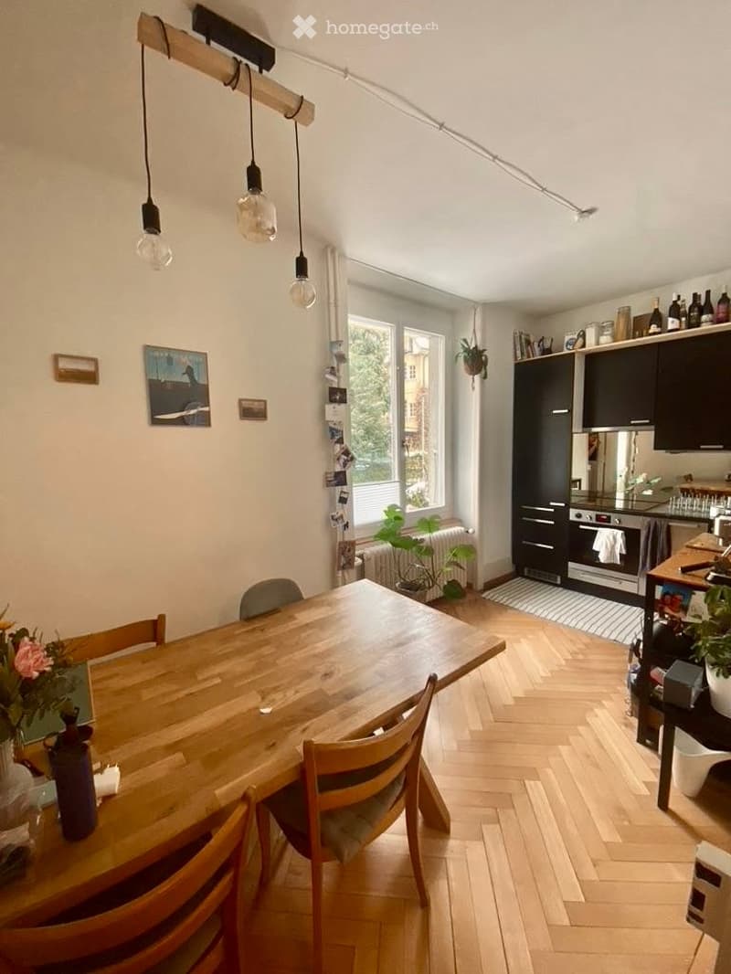 Open-plan kitchen and dining area with wooden flooring, a large wooden dining table with chairs, modern kitchen cabinets, and pendant lights.