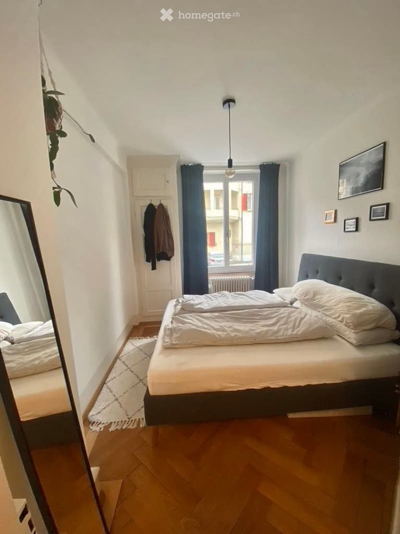 Cozy bedroom with wooden flooring, a large bed with white bedding, a mirror, and picture frames on the wall. A window with dark curtains lets in natural light.
