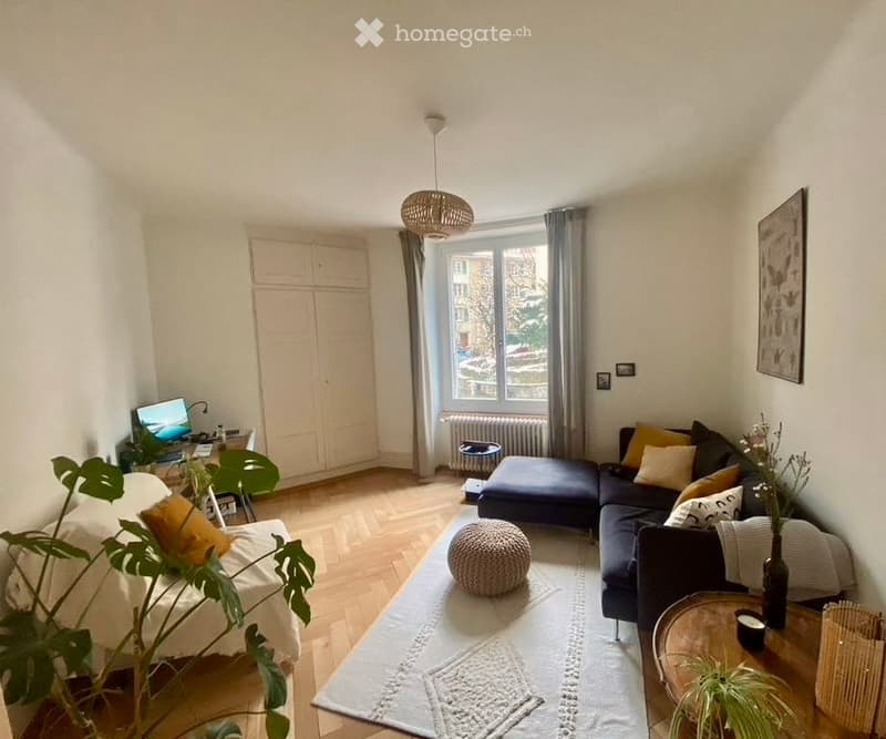 Modern living room with a sectional sofa, coffee table, ottoman, and a laptop on a desk. The room features wooden flooring, large windows with curtains, and decorative plants.