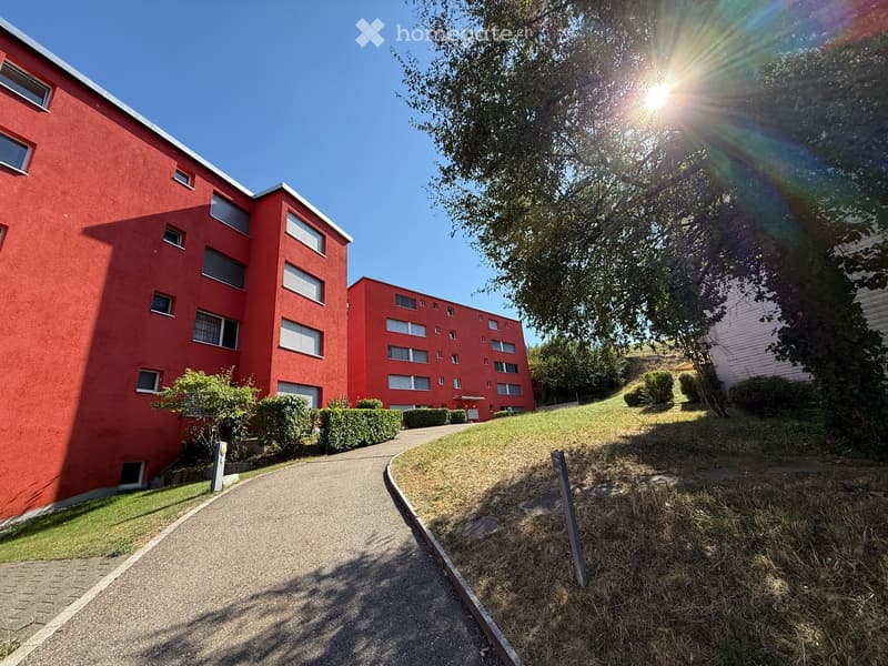 Red apartment building with multiple windows and balconies, surrounded by greenery and a paved pathway.