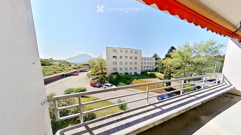 Balcony with metal railing overlooking a parking lot, buildings, and greenery. Red and yellow awning visible at the edge of the balcony.