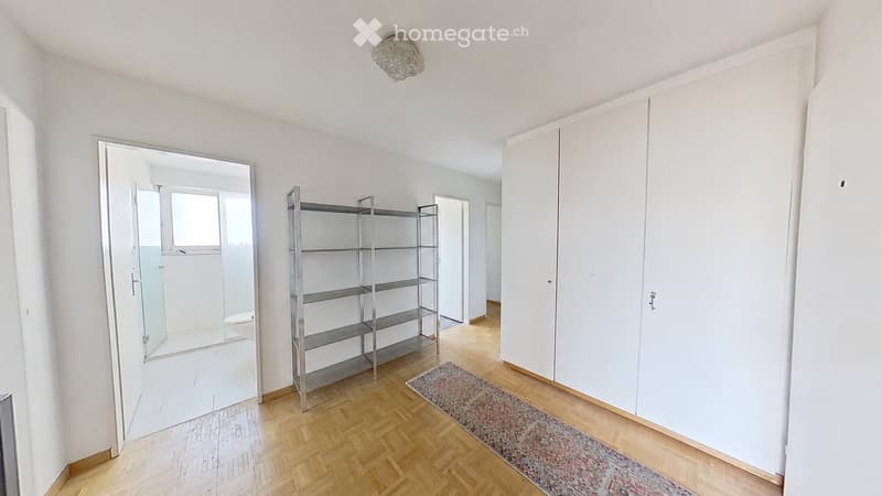 Bright, minimalist room with wooden flooring, white walls, a large built-in wardrobe, a metal shelving unit, a large mirror, and a patterned rug.