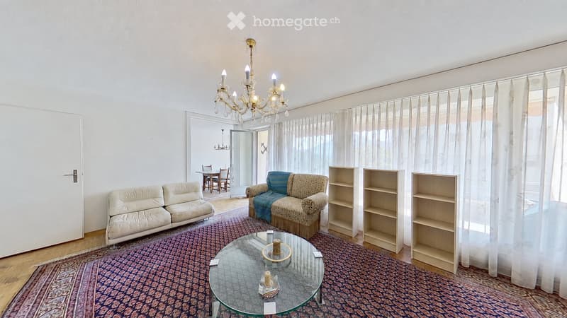 Living room with a white leather sofa, patterned armchair, glass coffee table, and shelves. A dining area is visible through an open doorway.