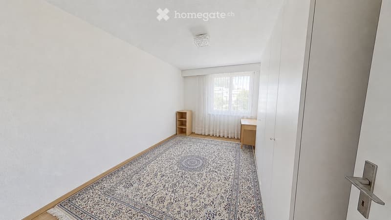A small, unfurnished room with white walls, wooden flooring, a patterned rug, a small shelf, and a small table near the window.