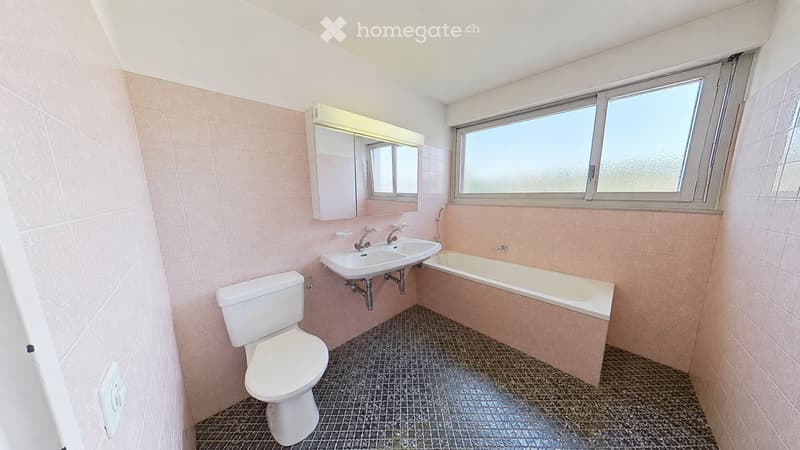 Bathroom with pink tiles, a toilet, a double sink, a bathtub, and a mirror above the sink. The room has a window with frosted glass and a dark tiled floor.