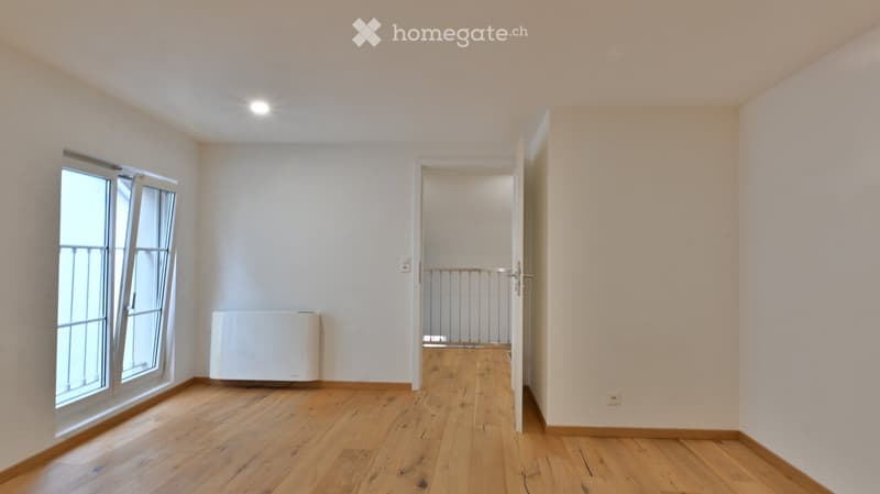 Empty room with wooden flooring, white walls, and a large window with a view of a balcony. A radiator is visible against one wall, and an open doorway leads to another room with a staircase.