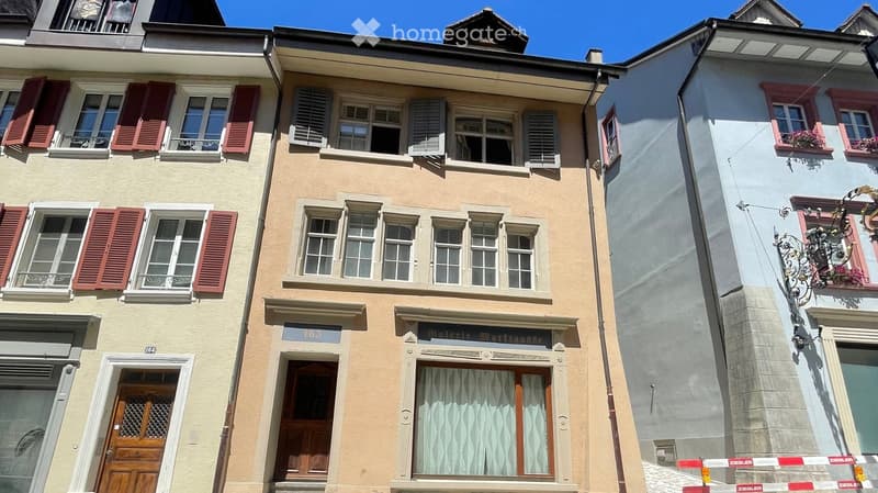 Three-story building with multiple windows, shutters, and balconies. The central building has a sign reading "Galerie Martreuse" and a curtained window on the ground floor.