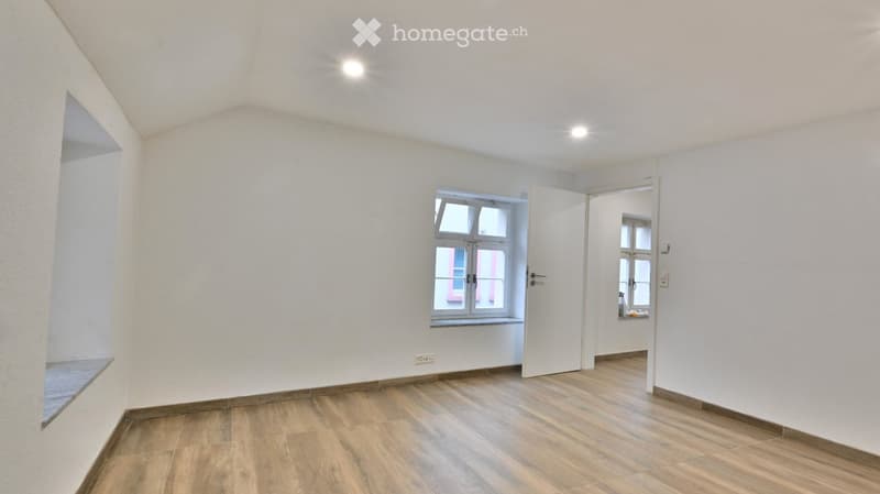 Empty room with wooden flooring, white walls, and a window with a view of a red building. The room has a door leading to another space.