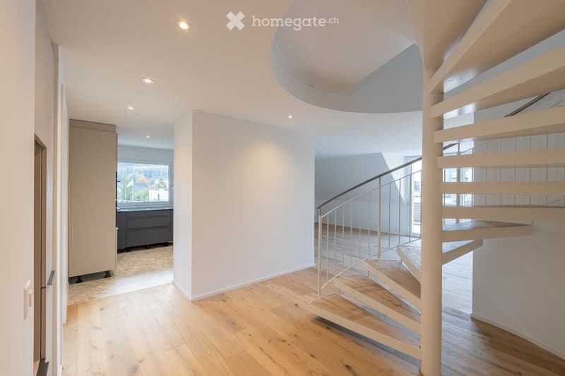 Modern hallway with wooden flooring, a curved staircase with a glass railing, recessed ceiling lights, and a view of a room with a window and cabinetry.