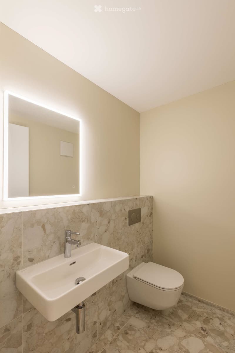 Modern bathroom with a white sink, chrome faucet, toilet, and large illuminated mirror above the sink. The walls are tiled with a stone-like pattern, and the floor has a similar stone tile design.