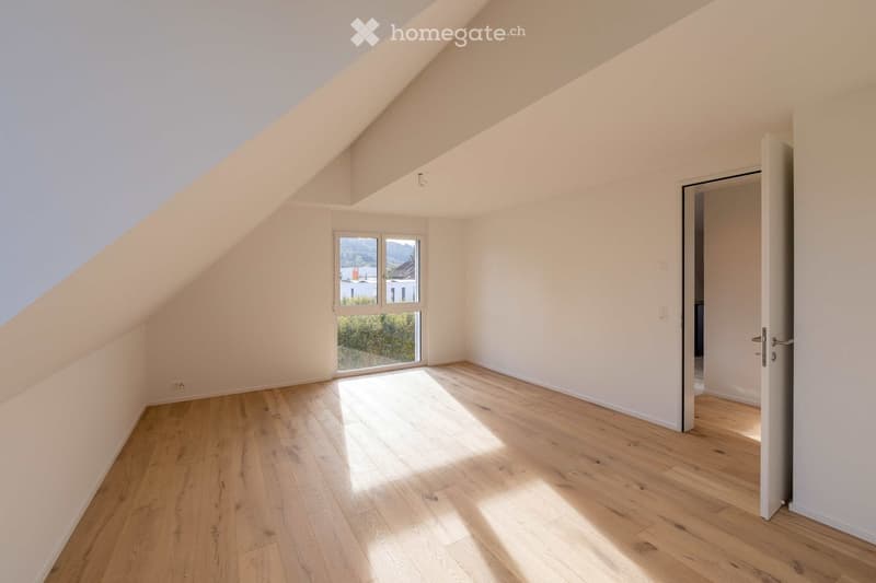 Bright, empty attic room with wooden flooring, white walls, and large windows letting in natural light. The room features a sloped ceiling and an open doorway leading to another space.