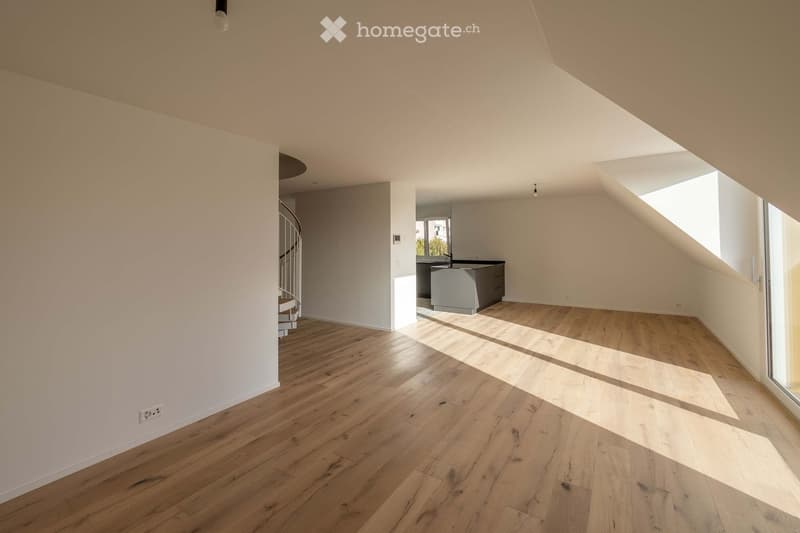 Bright, empty room with wooden flooring and white walls, featuring a spiral staircase leading to another level and a view of a modern kitchen area in the background.