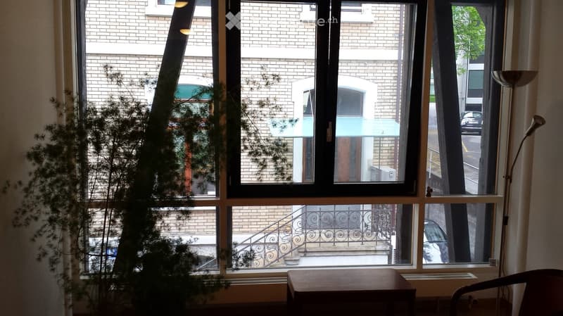 A room with large windows overlooking a brick building and a staircase. Inside, there is a wooden table, a chair, and a floor lamp near the window.