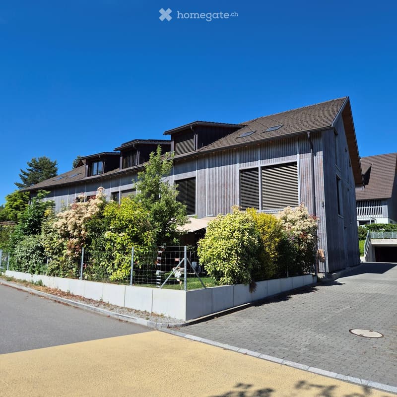 Modern two-story house with wooden exterior, multiple windows, and a fenced garden area.