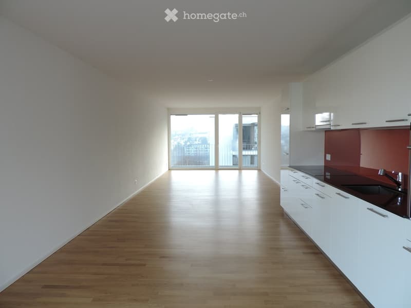 Empty room with wooden flooring, white walls, and a small kitchen area with white cabinets and a red backsplash.