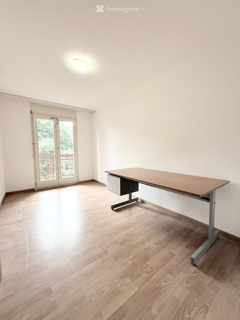 A room with light wooden flooring, white walls, and a large sliding glass door leading to a balcony. A single wooden table with drawers is positioned against the wall.