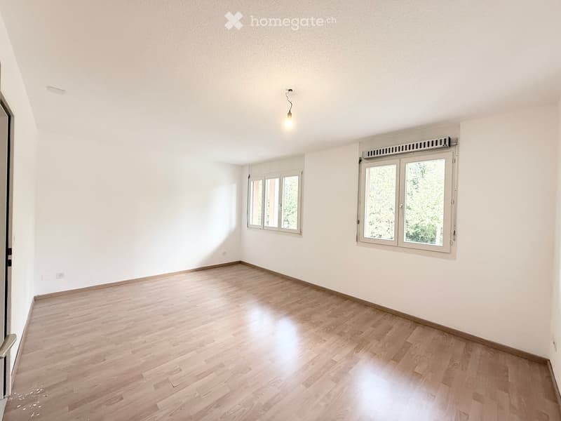 Empty room with light wooden flooring, white walls, and large windows allowing natural light. The room features a single hanging light fixture and a ventilation unit above one of the windows.