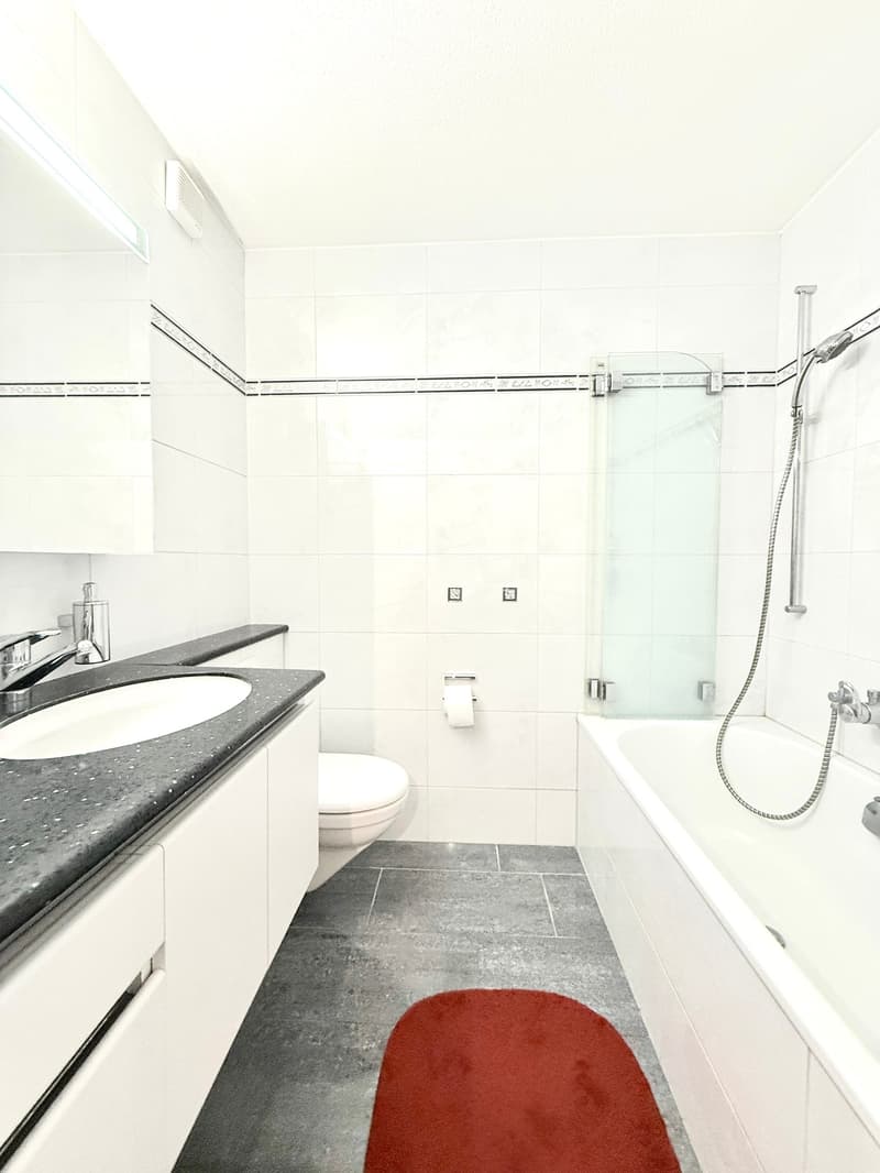 Bathroom. Modern bathroom with white tiles, a black countertop sink, a toilet, a bathtub with a glass shower enclosure, and a red bath mat on the floor.