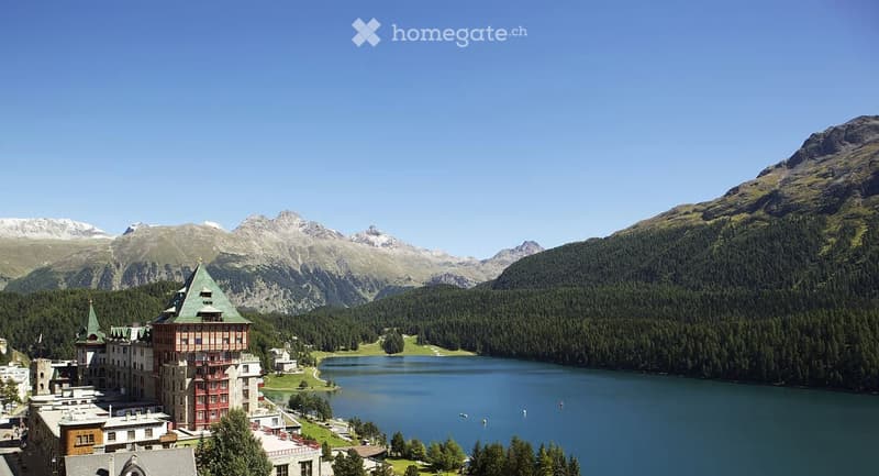 A large, historic building with a green roof and red accents, situated near a serene lake surrounded by lush forests and towering mountains.