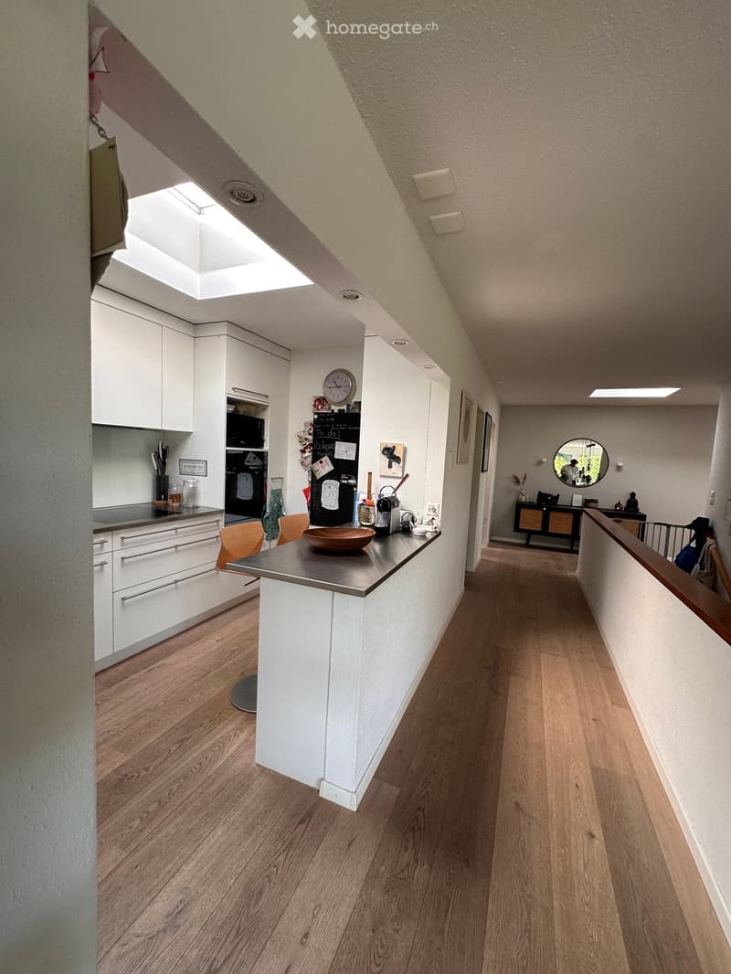 Bright hallway with wooden flooring, white walls, and a modern kitchen area visible on the left side. The kitchen features white cabinets, a stainless steel island, and various appliances.