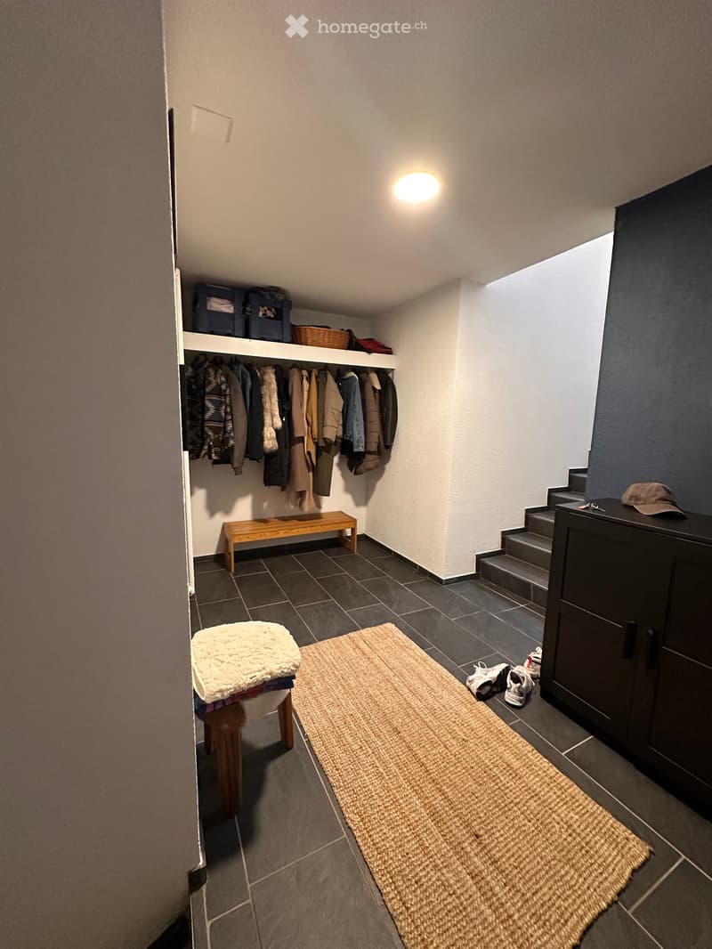 A narrow hallway with tiled flooring, a built-in coat rack, a small wooden bench, a woven rug, and a staircase leading upwards.