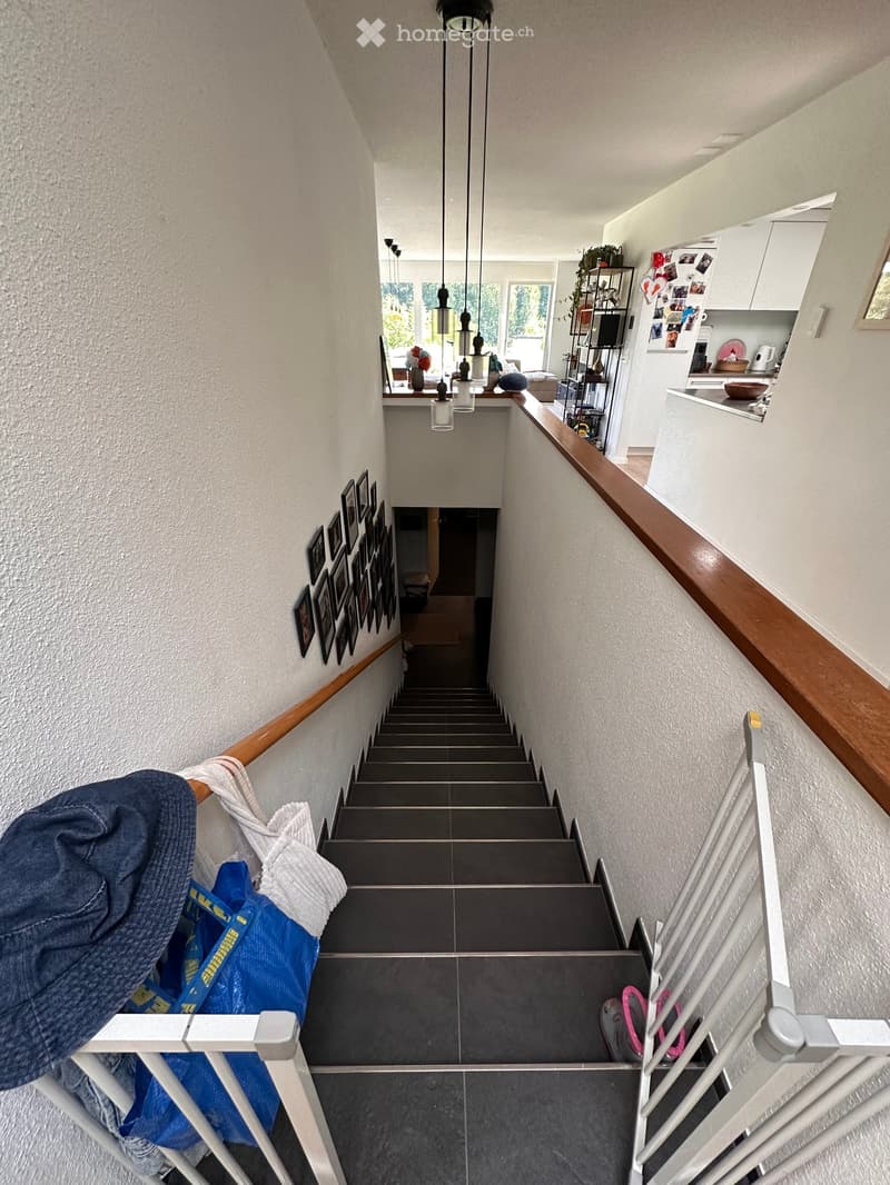 Staircase with wooden handrails, grey tiled steps, and a white baby gate at the bottom. The walls are painted white and decorated with framed pictures. A blue bag and a folded towel are placed on the left side of the stairs.