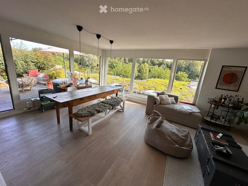 Bright living room with large windows offering a scenic view, featuring a sofa, coffee table, dining table, chairs, a bench with a fur throw, a beanie bag, and a bar cart.
