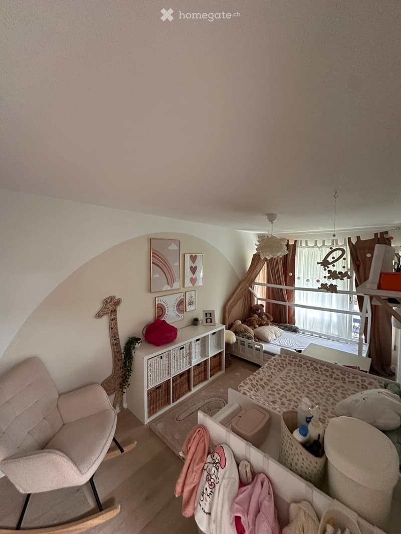 A cozy children's bedroom with a slanted ceiling, featuring a white shelf with storage baskets, a pink rocking chair, a small bed with stuffed animals, and a changing table with baby items.