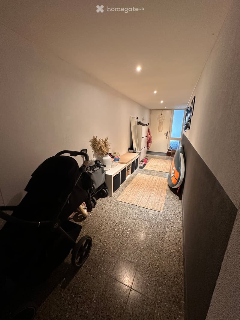 Narrow hallway with tiled floor, small bench with storage, baby stroller, and decorative items. Light-colored walls and recessed ceiling lighting.