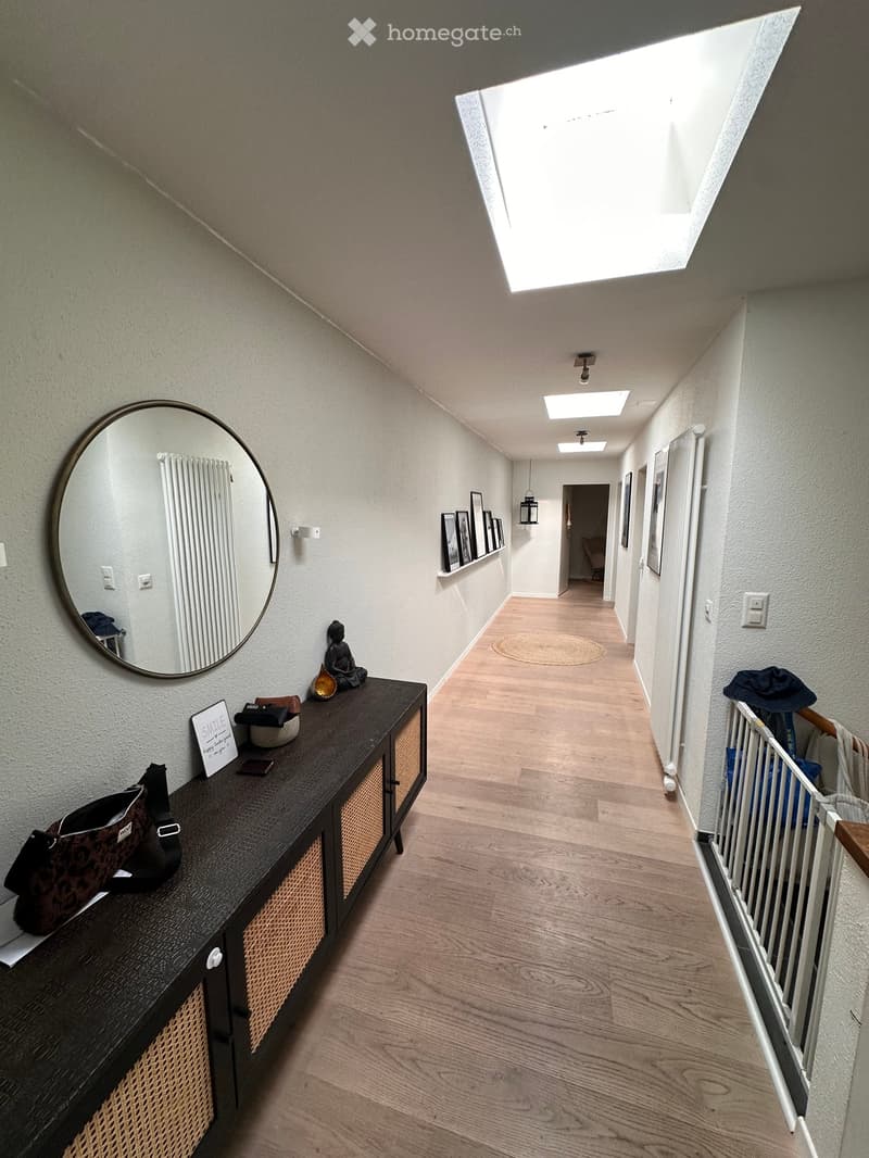 A modern hallway with wooden flooring, white walls, and a skylight at the top. Features include a dark wooden console table with a mirror, decorative items, and framed pictures on the walls.