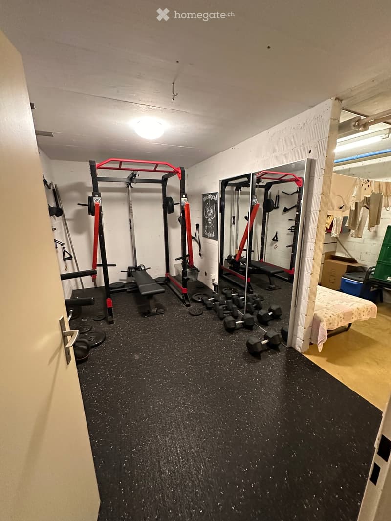A well-equipped home gym with a weight bench, dumbbells, weight rack, and a mirror. The space features a black rubber floor and white walls.