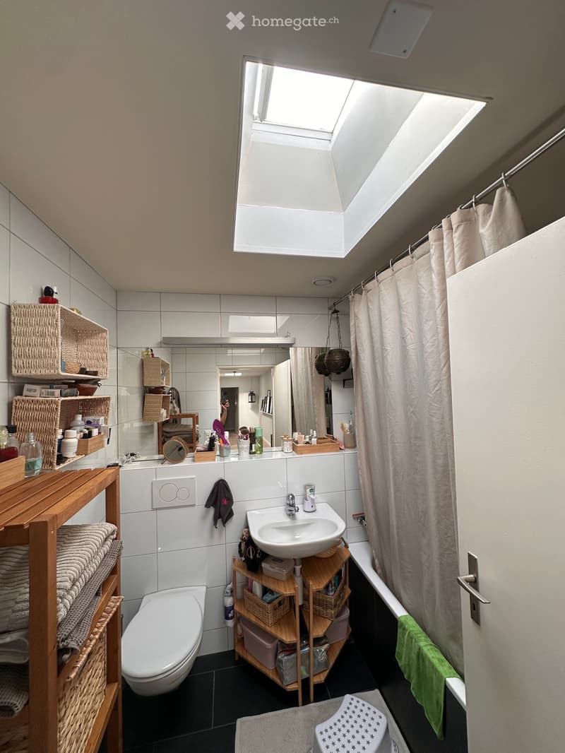 Modern bathroom with white tiles, a skylight window, a toilet, a sink with a mirror, a shower area with a curtain, and wooden shelves holding towels and toiletries.