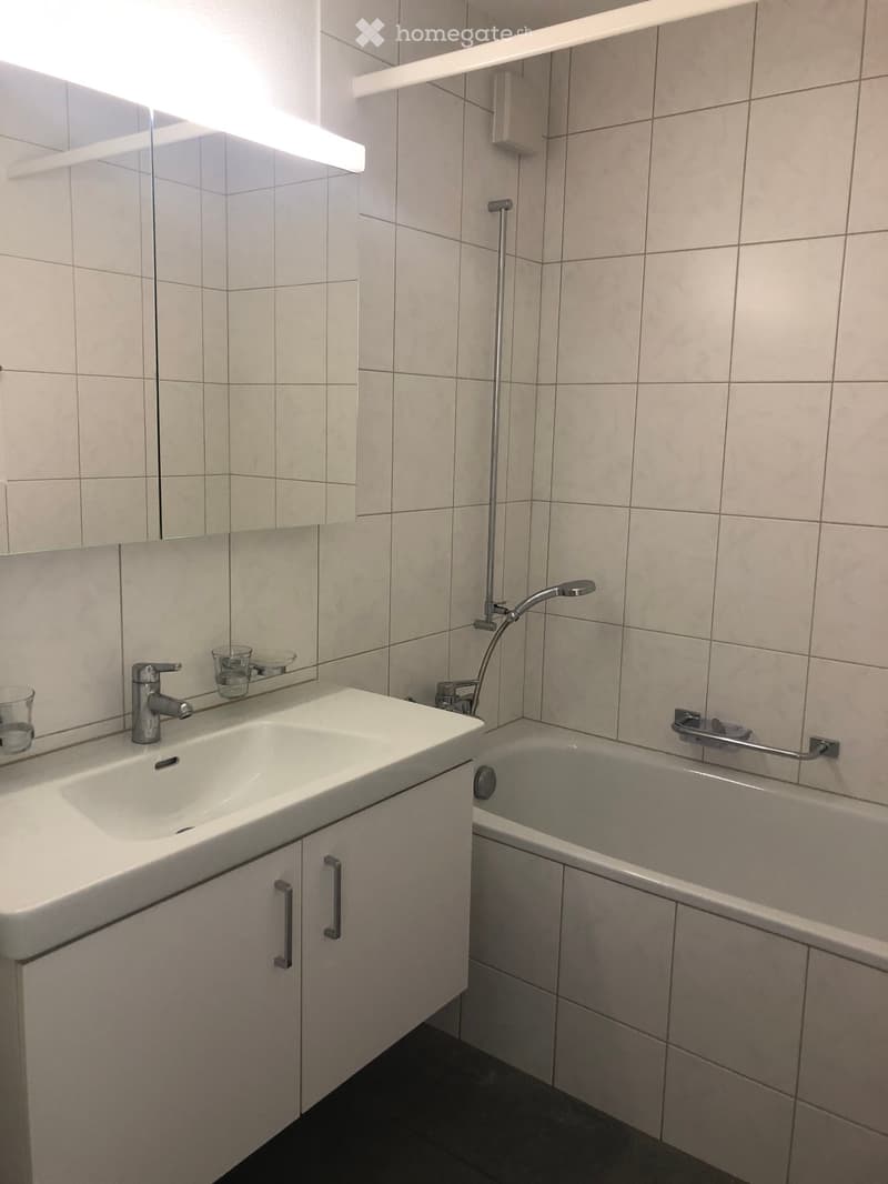 Modern bathroom with a white sink, faucet, bathtub, and mirror. The walls are tiled in light beige, and the floor is dark.