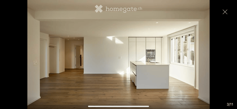 Modern kitchen with white cabinets, a central island, and large windows allowing natural light to fill the space. The floor is made of polished wood.