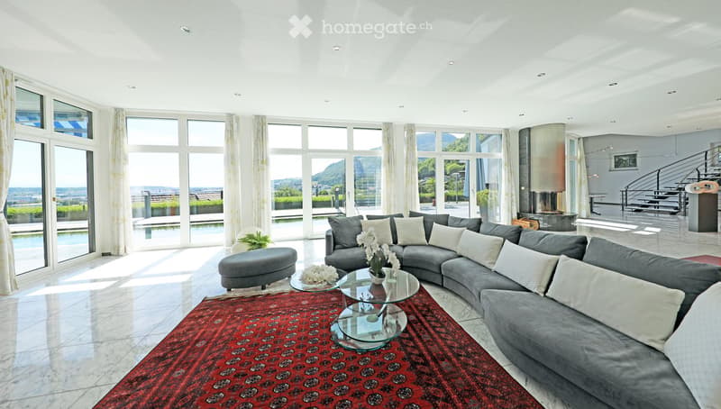 Modern living room with large windows offering a scenic view, featuring a grey sectional sofa, glass coffee tables, an ottoman, and a red patterned rug.