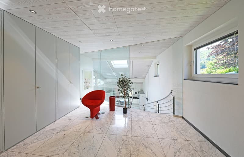 Modern hallway with marble flooring, glass walls, a red chair, a potted plant, and a trash can. The hallway features a staircase with a metal railing and a large window offering a view of greenery.