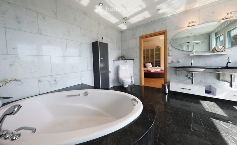 Modern bathroom with a large bathtub, toilet, double sink, and a large oval mirror. The room features marble tiles and a black polished floor.