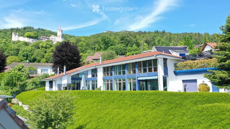 Modern two-story house with large windows, red-tiled roof, and a blue awning, situated on a hillside with a well-maintained lawn and surrounding greenery.