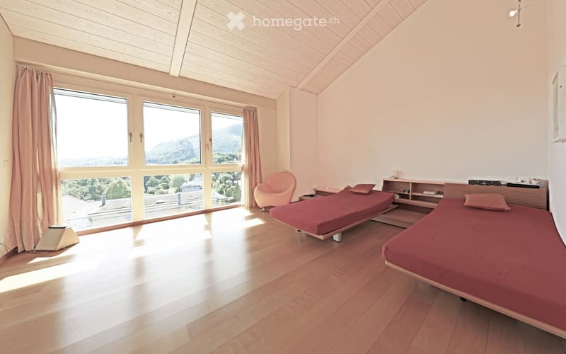 Bright bedroom with wooden flooring, two beds with red bedding, a pink armchair, and large windows offering a scenic view.