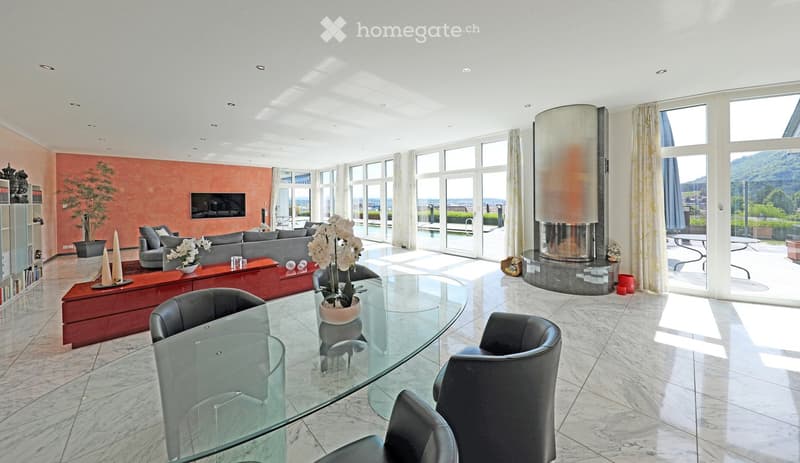 Modern living room with large windows offering a scenic view, featuring a grey sofa, glass dining table with black chairs, a central fireplace, and a television mounted on a vibrant orange accent wall.