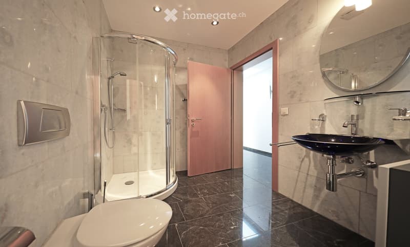 Modern bathroom with marble walls, a glass-enclosed shower, a toilet, a sink with a modern faucet, and a large round mirror.