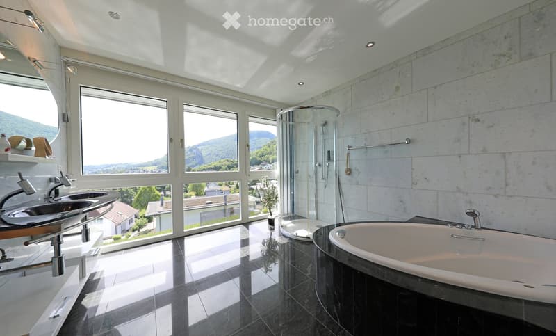 Modern bathroom with large windows offering a scenic view, featuring a bathtub, double sink, and a glass-enclosed shower.