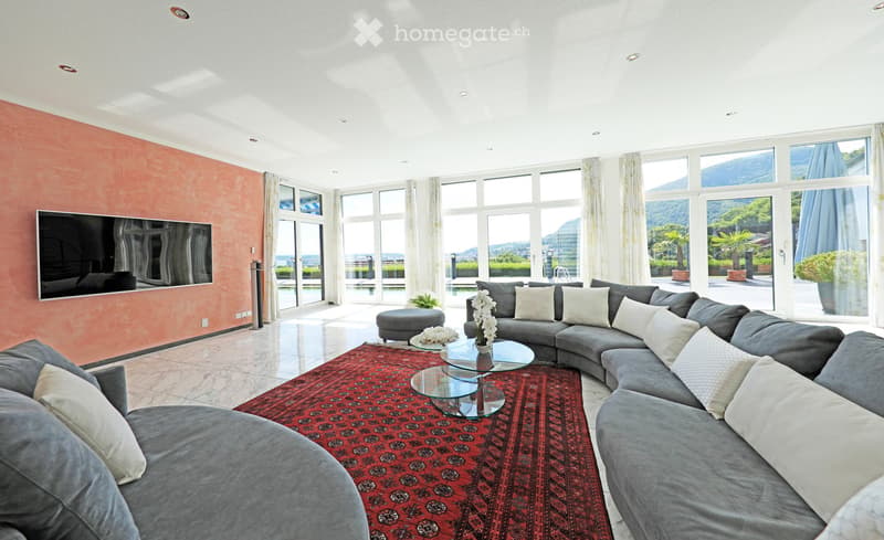 Modern living room with large windows offering a scenic view, grey sectional sofa, coffee tables, ottoman, television, and decorative flower vases.