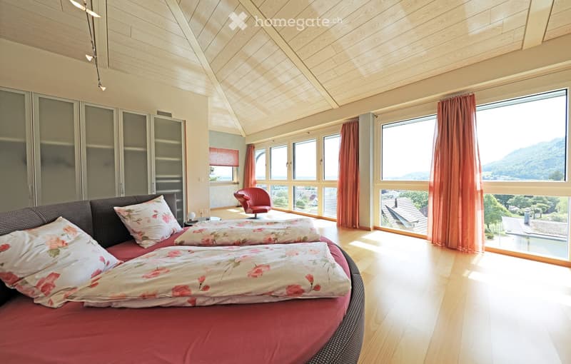 Bright bedroom with wooden floors, large windows offering a scenic mountain view, a round bed with floral bedding, a red armchair, and a modern wardrobe.