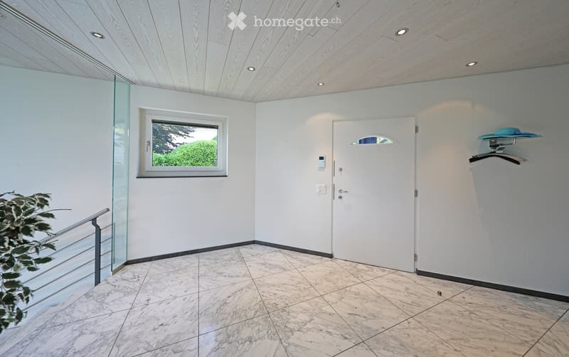 Modern hallway with white walls, marble flooring, recessed lighting, a large window, and a coat rack with a hat on it.