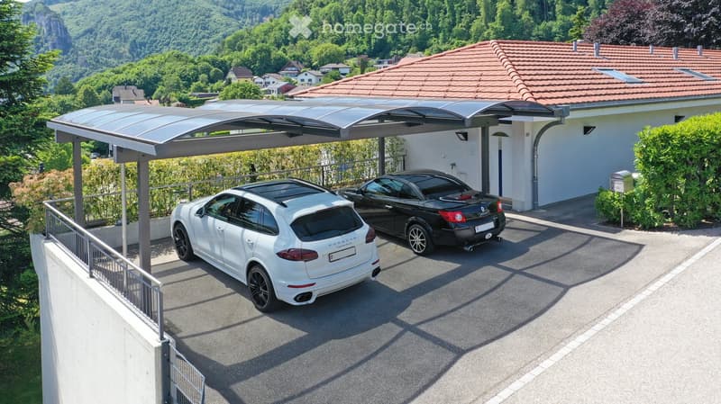 Two cars parked under a modern carport attached to a house, with a scenic mountainous background.